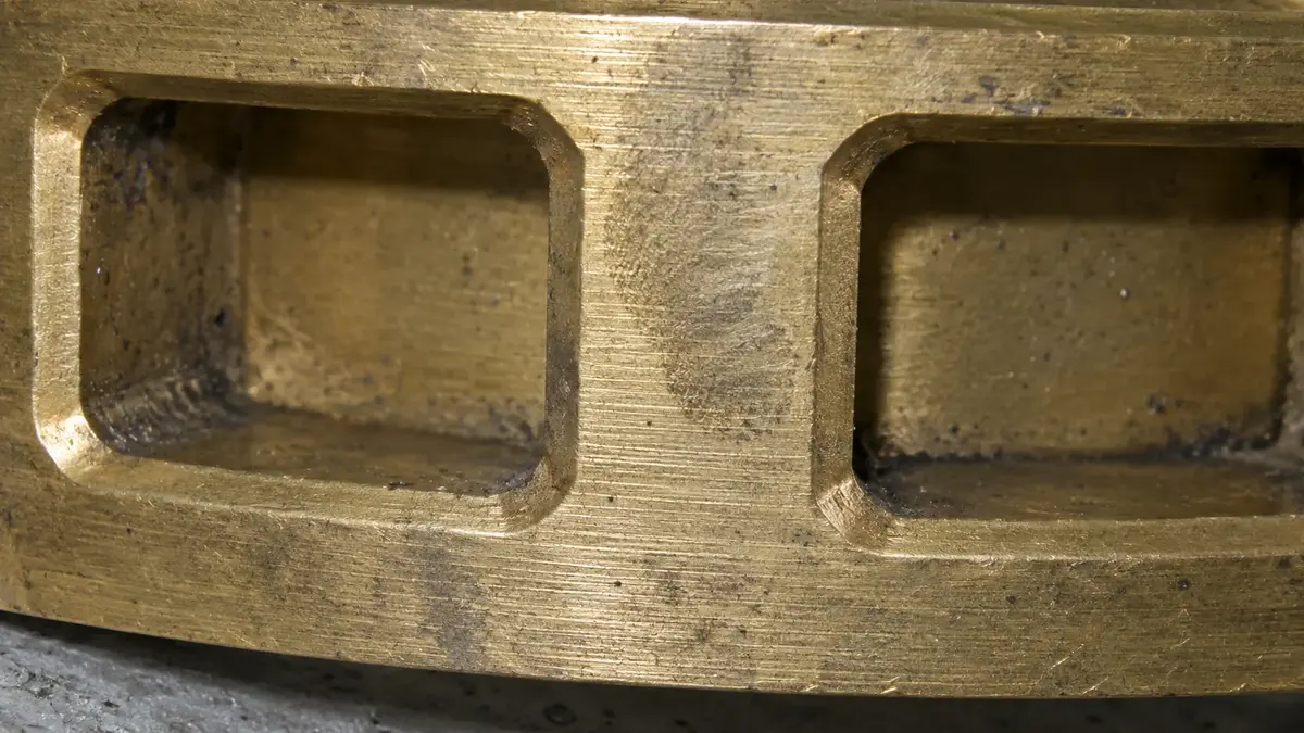 Two window pockets in a machined brass bearing cage at close range, with a chamfer worked around each opening and the thick solid wall visible through the window, the features that separate a machined cage from a stamped sheet one