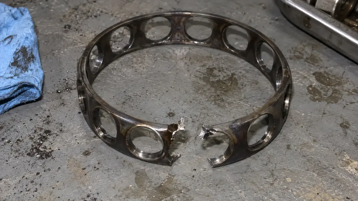 A steel bearing cage with one bar torn completely through and the torn ends bent outward, the fresh fracture faces bright against the brown heat tint carried by the intact bars, the pattern left when cage damage follows a mounting or lubrication fault rather than a weak cage