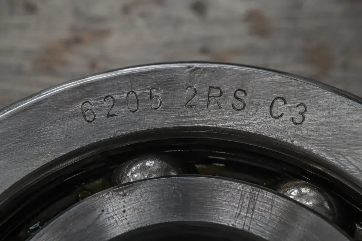 Macro close-up of a deep groove ball bearing outer ring laser-etched with the full designation 6205 2RS C3, with the rolling elements visible below the marking