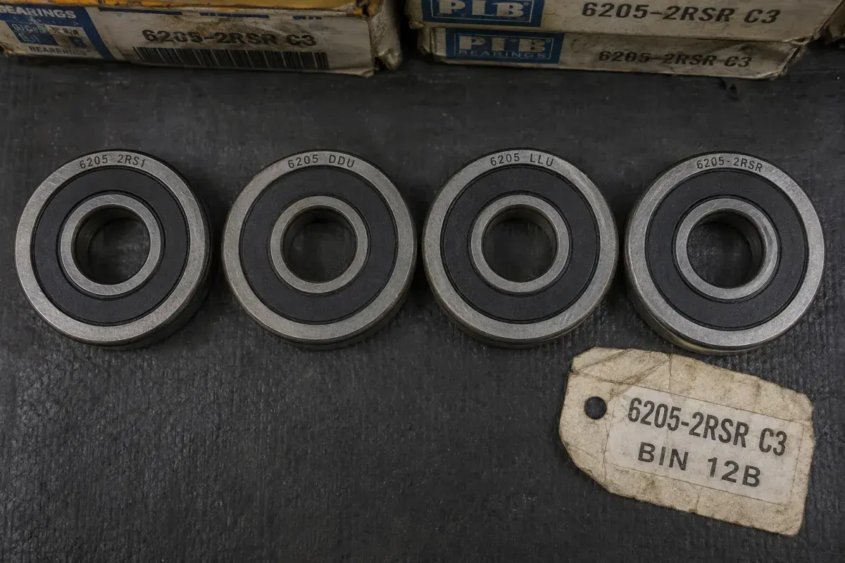 Four size-identical 6205 bearings in a row, each with matte black rubber contact seals but a different brand suffix stamped on the ring — 6205-2RS1, 6205 DDU, 6205 LLU, and 6205-2RSR — beside a warehouse bin tag