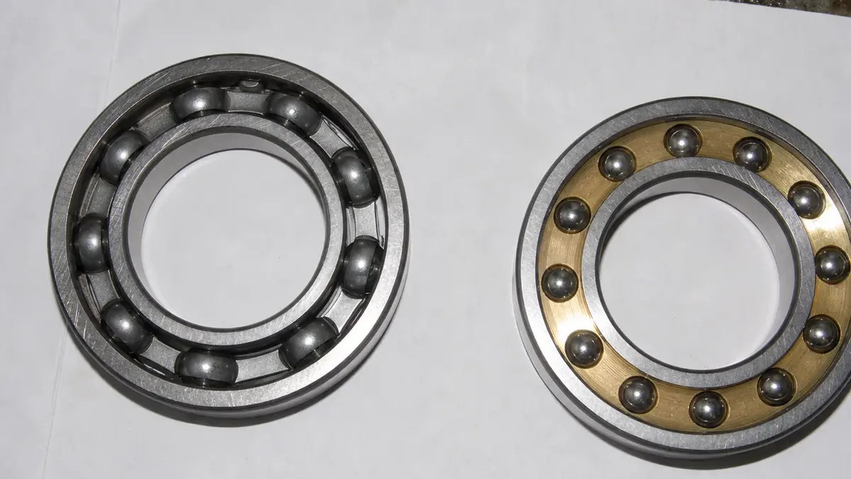 Two open ball bearings compared side by side, the left with a stamped steel cage and the right with a machined brass cage, the cage type being a separate control item in a low-noise bearing specification