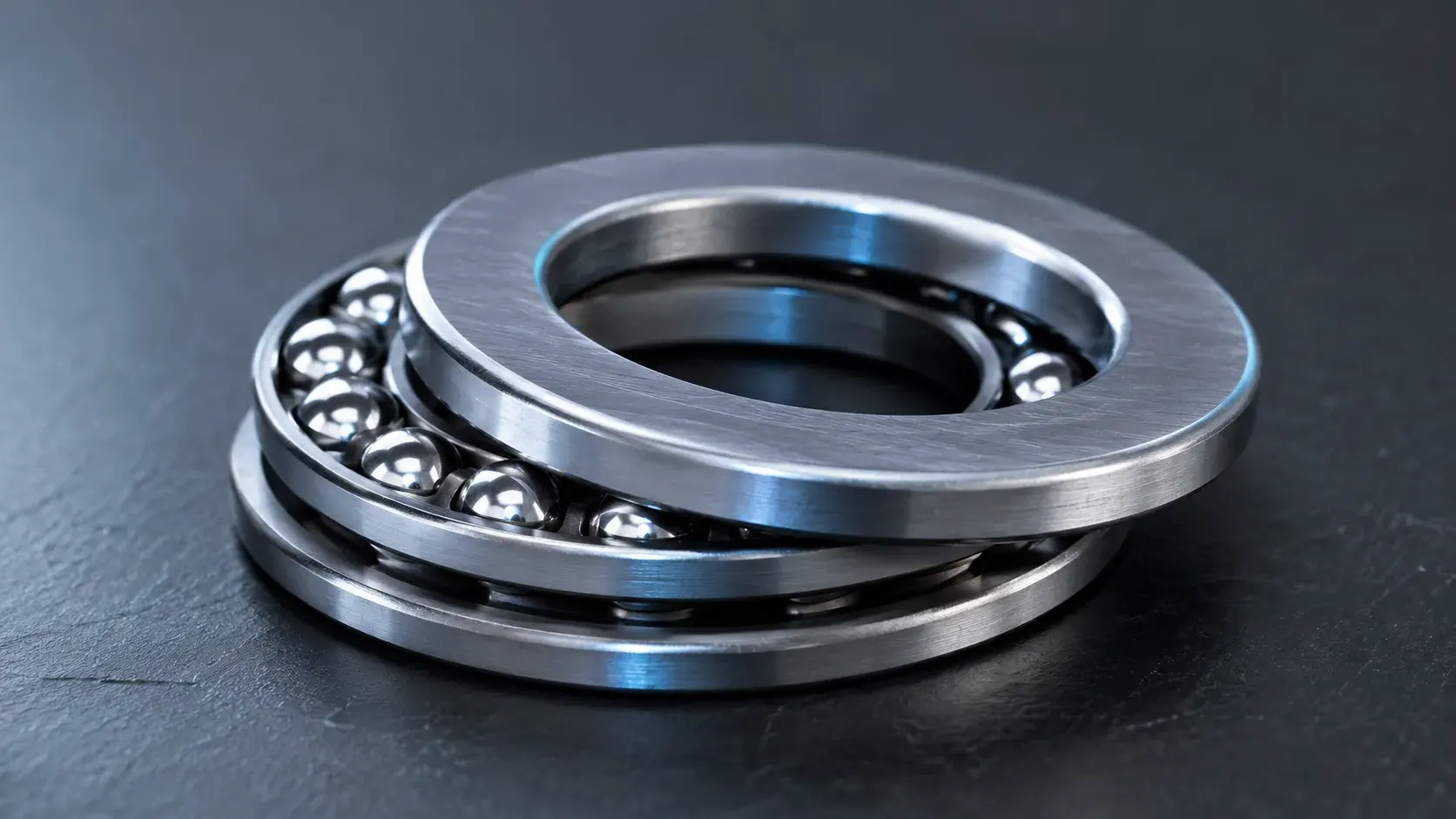 Single-direction thrust ball bearing on dark slate, its two flat washers separated to reveal the row of steel balls and the cage between them