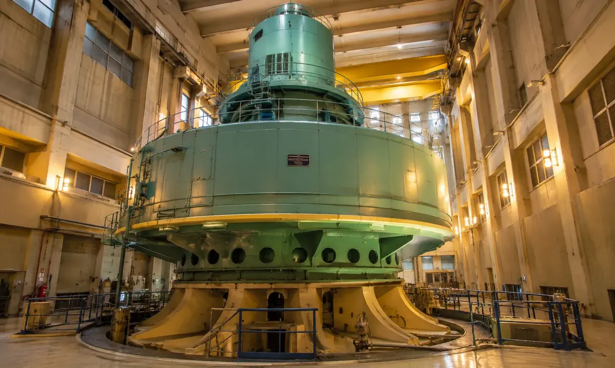 Interior of a hydroelectric power plant with a massive vertical-shaft generator turbine dwarfing the surrounding walkways and platforms